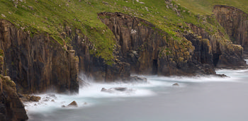Long exposure Cornish cliffs two This landscape photograph captures the Cornish cliffs two along the coastline in Cornwall during the late afternoon in early summer. The long exposure technique emphasizes the motion of the sea as it washes around the base of the steep cliffs, creating a smooth, misty effect on the water. The cliffs are rugged and partially covered in green vegetation, with visible rock formations and several caves carved into their faces. The image showcases the natural beauty of Cornwall, highlighting the interaction between the sea and the imposing cliffs, and the presence of caves that are typical features in the coastal geology of the region.